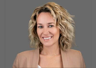 A person with wavy blonde hair and a beige blazer smiles in front of a grey background.