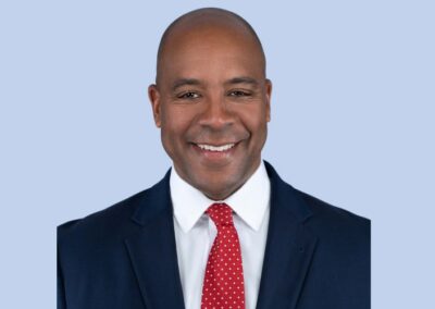 A person in a dark blue suit, white shirt, and red polka dot tie, smiling against a light blue background.