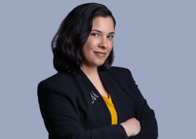 A woman with shoulder-length dark hair, wearing a black blazer and yellow shirt, stands with her arms crossed against a light blue background.