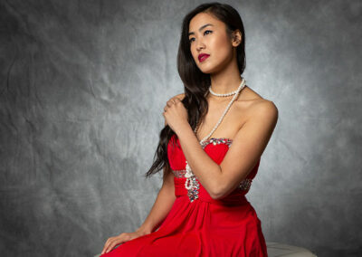 A woman in a red strapless dress with a pearl necklace poses against a textured gray background.