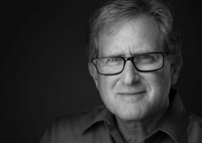 A grayscale close-up photo of a middle-aged man with glasses, short hair, and a slight smile, wearing a collared shirt, against a dark background.