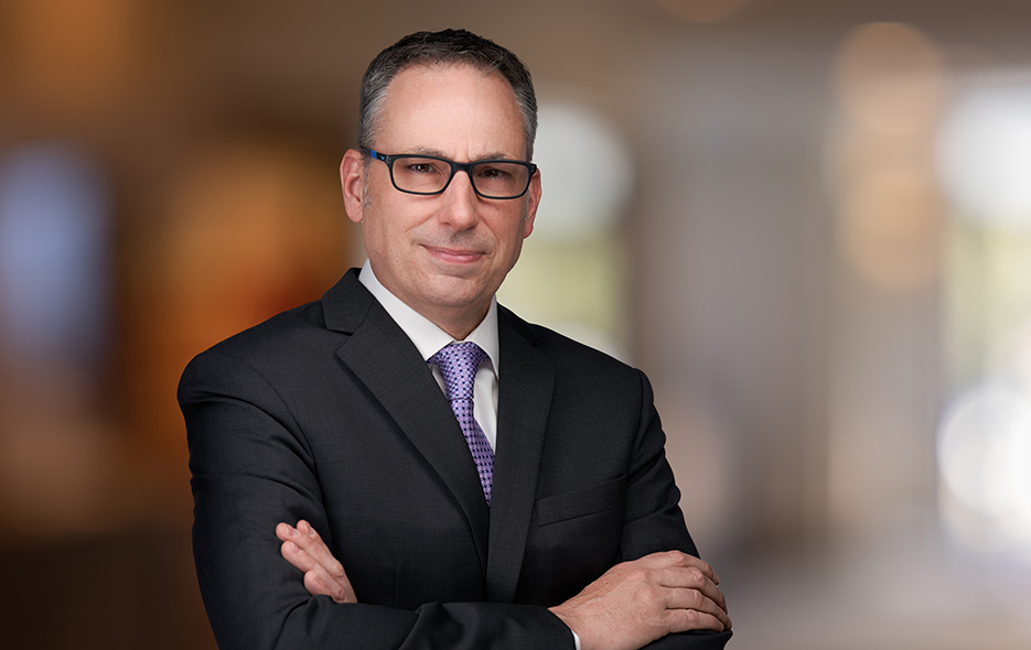 Middle aged white male, brown hair and glasses wearing a dark suit, tie and white shirt.  Posed for a professional photo shoot.