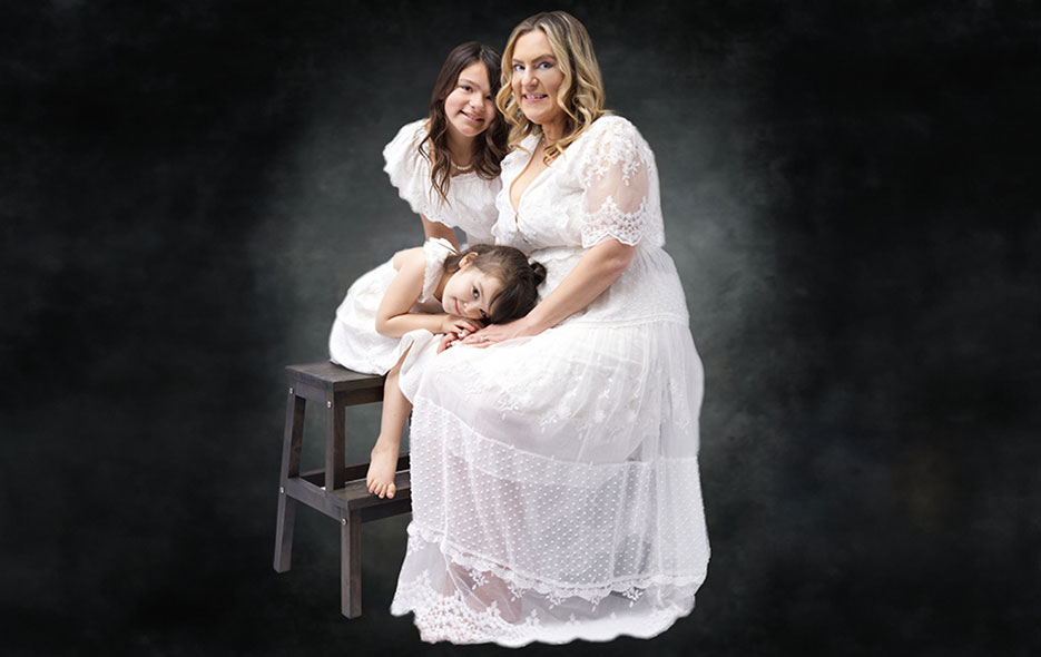 Mom and 2 young daughters posed on a stool in white dresses.