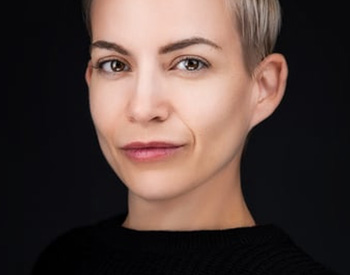 Person with short, light-colored hair, wearing a dark top, against a dark background.