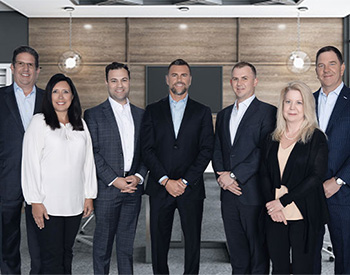 A group of seven people in business attire stand together in an office setting.