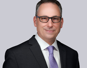 A person in a suit, wearing glasses, poses against a gray background.