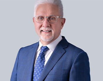 A man with short white hair, glasses, and a beard wearing a blue checkered suit and patterned tie, standing against a light gray background.