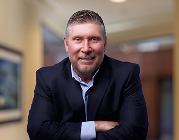 A man in a dark suit and light blue shirt smiles, leaning forward with arms crossed in an office setting.